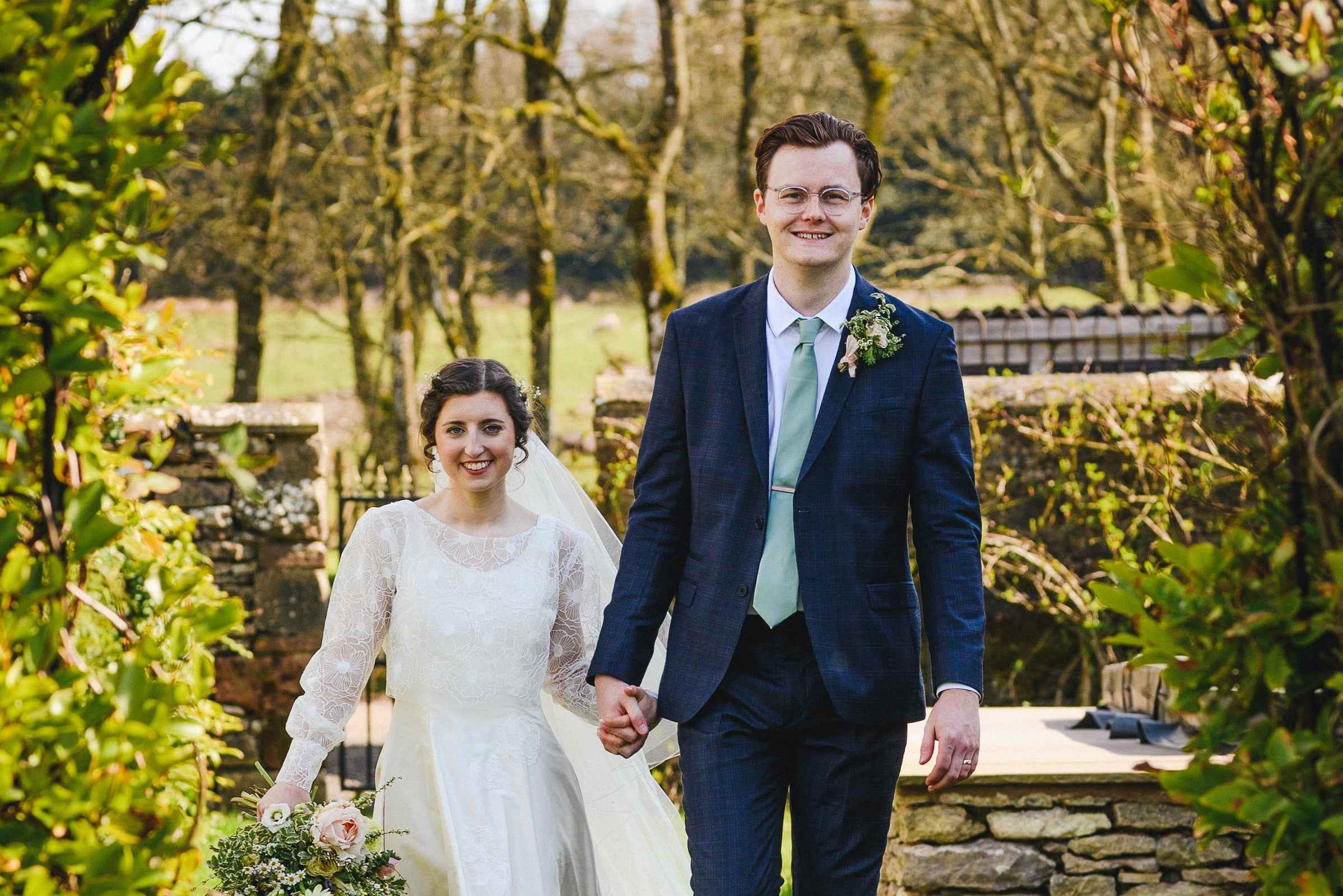 Lovio founders on their wedding day, holding hands outdoors with greenery and stone wall in the background